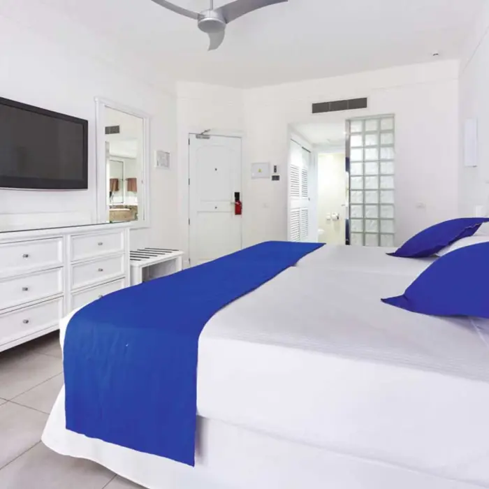 A clean, white connecting double standard room with two beds, blue accents, a TV, and a view into an adjacent room through an open doorway.