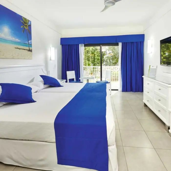 A bright standard room with two beds adorned with blue accents, a flat-screen TV, and a balcony with lush greenery visible through the open curtains.