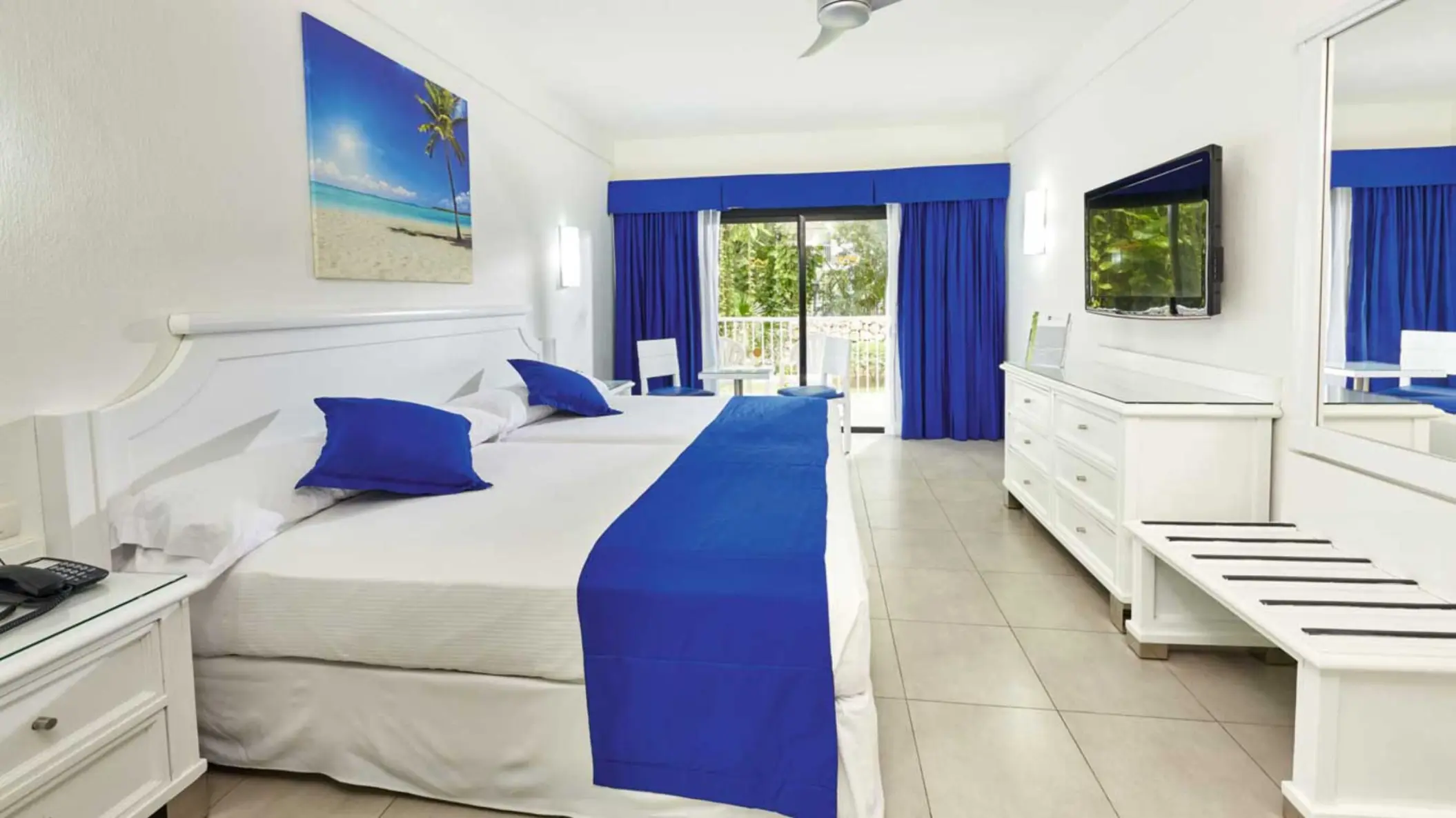 A bright standard room with two beds adorned with blue accents, a flat-screen TV, and a balcony with lush greenery visible through the open curtains.