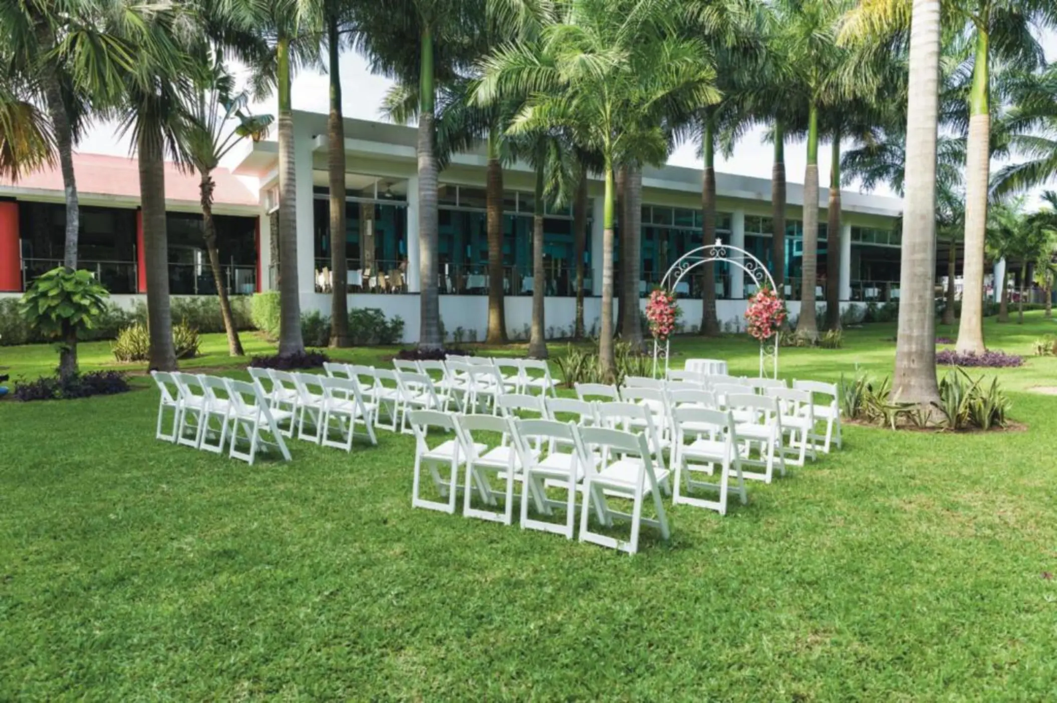 A serene outdoor garden ceremony setup on a lush green lawn, featuring rows of white folding chairs and a decorative arch.