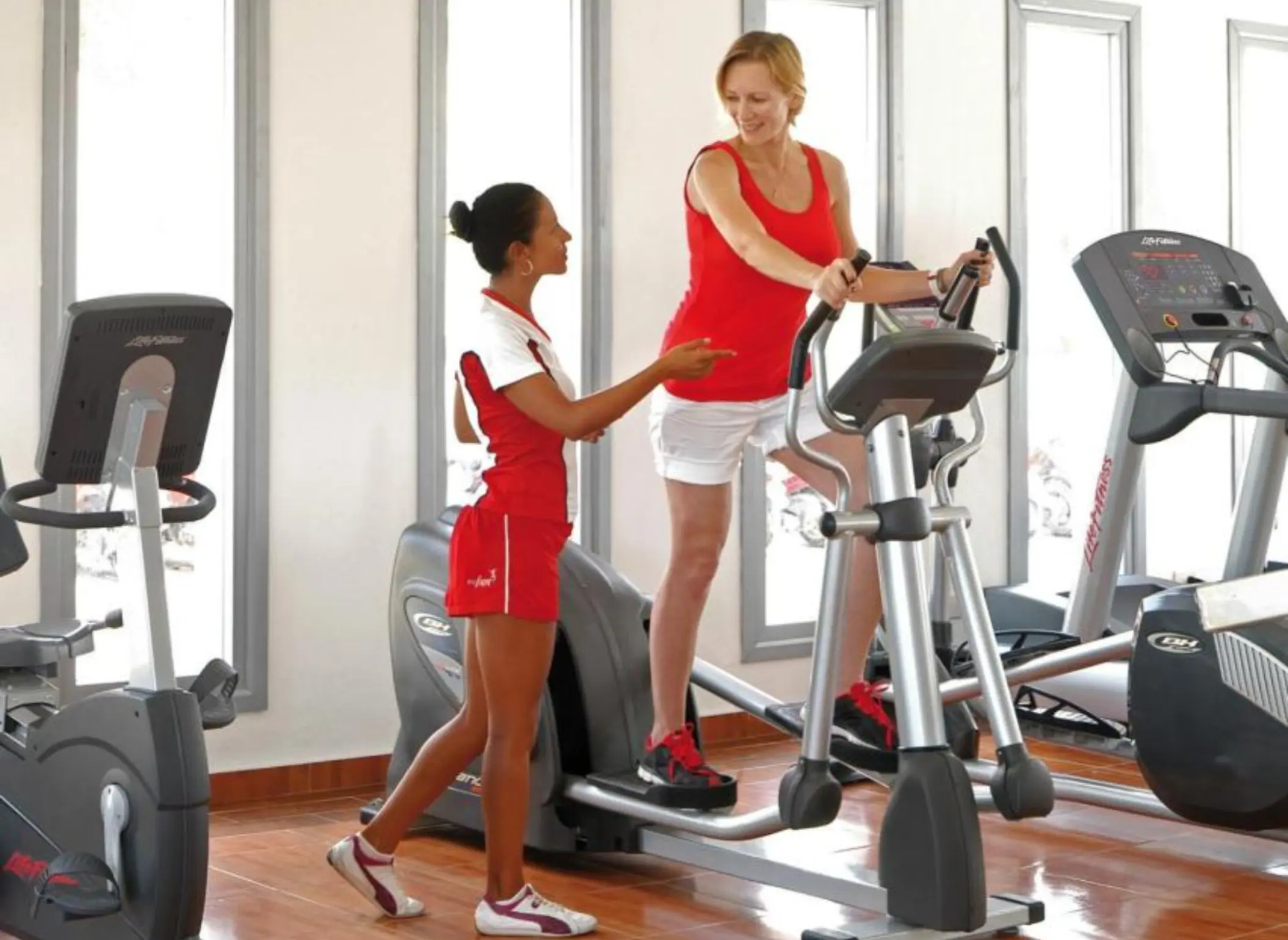 a bright gym with large windows; one on an elliptical machine, and the other assisting her.