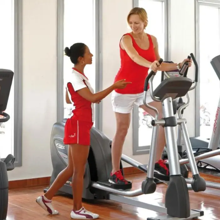 a bright gym with large windows; one on an elliptical machine, and the other assisting her.