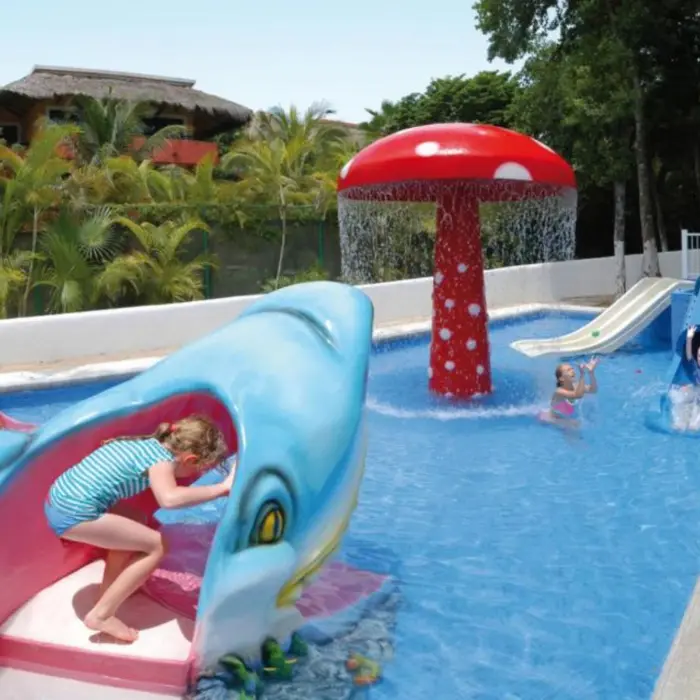 A vibrant kids' pool area featuring a mushroom-shaped water fountain and two water slides, one shaped like a shark, with children playing.