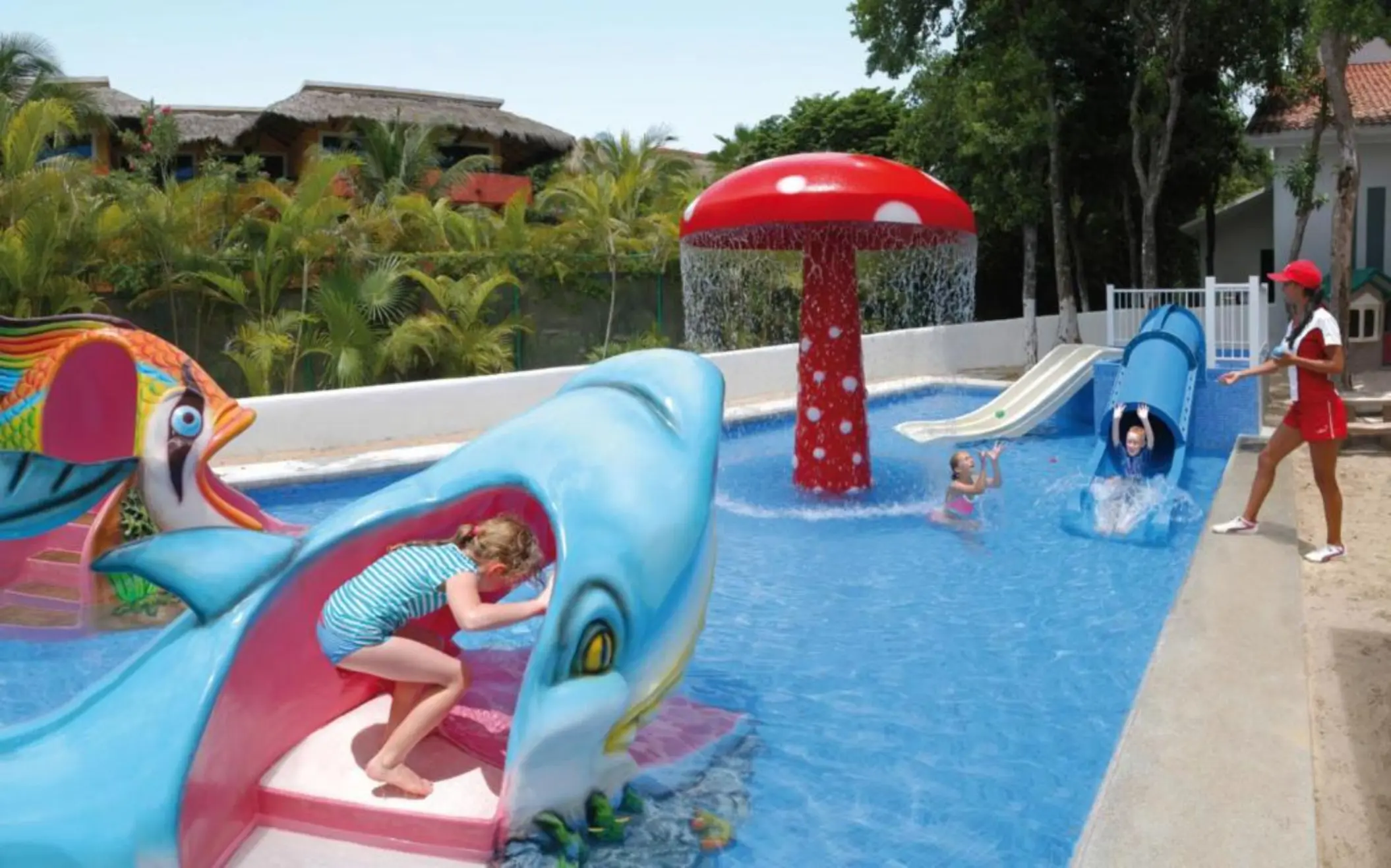 A vibrant kids' pool area featuring a mushroom-shaped water fountain and two water slides, one shaped like a shark, with children playing.