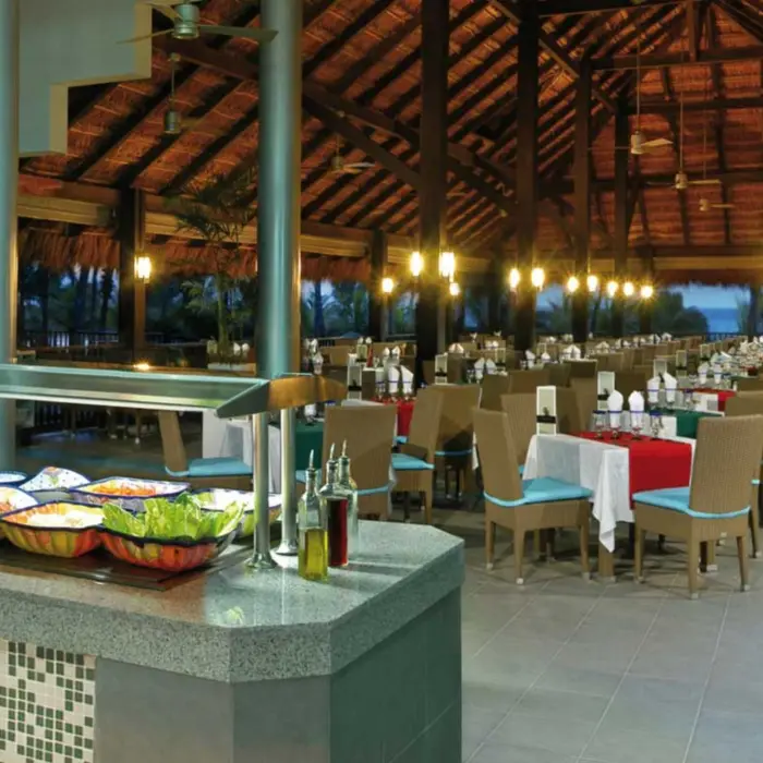 A large, open-air oceanview restaurant with a thatched roof, a buffet counter filled with fresh food, and numerous tables set for dining.