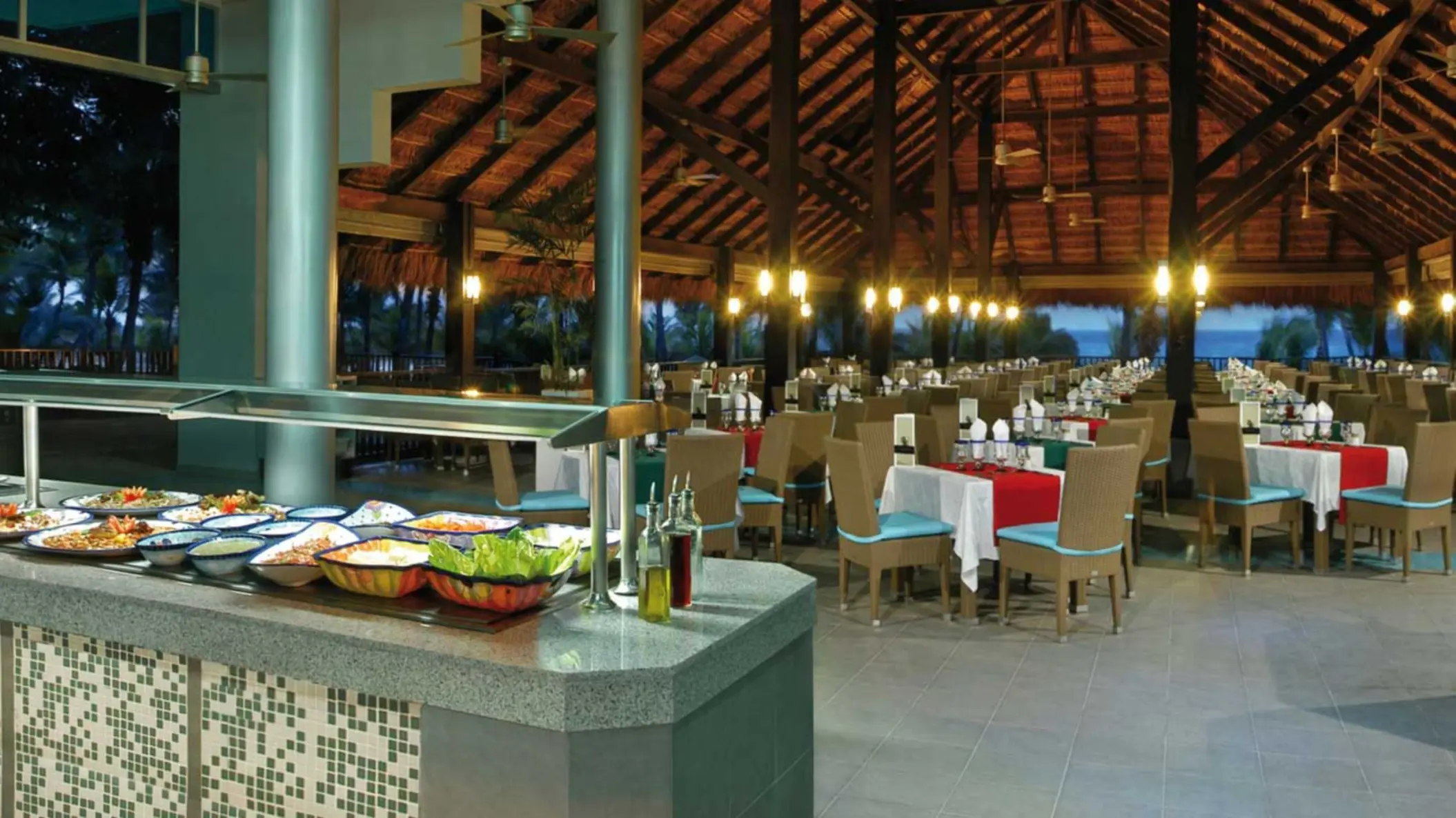A large, open-air oceanview restaurant with a thatched roof, a buffet counter filled with fresh food, and numerous tables set for dining.