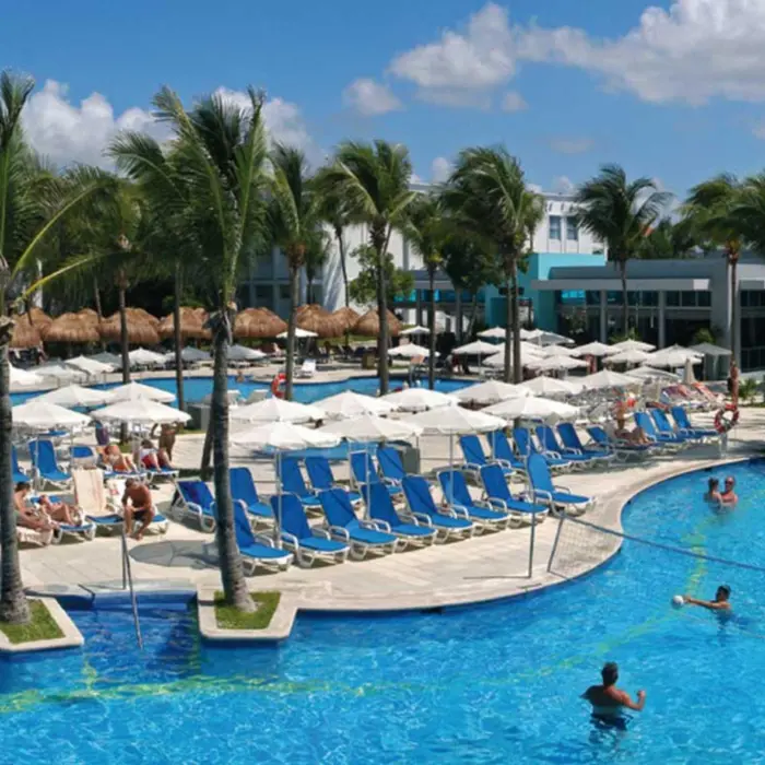 An expansive outdoor pool area at Hotel Riu Yucatan, featuring multiple interconnected pools, numerous lounge chairs, and palm trees under a bright sky.