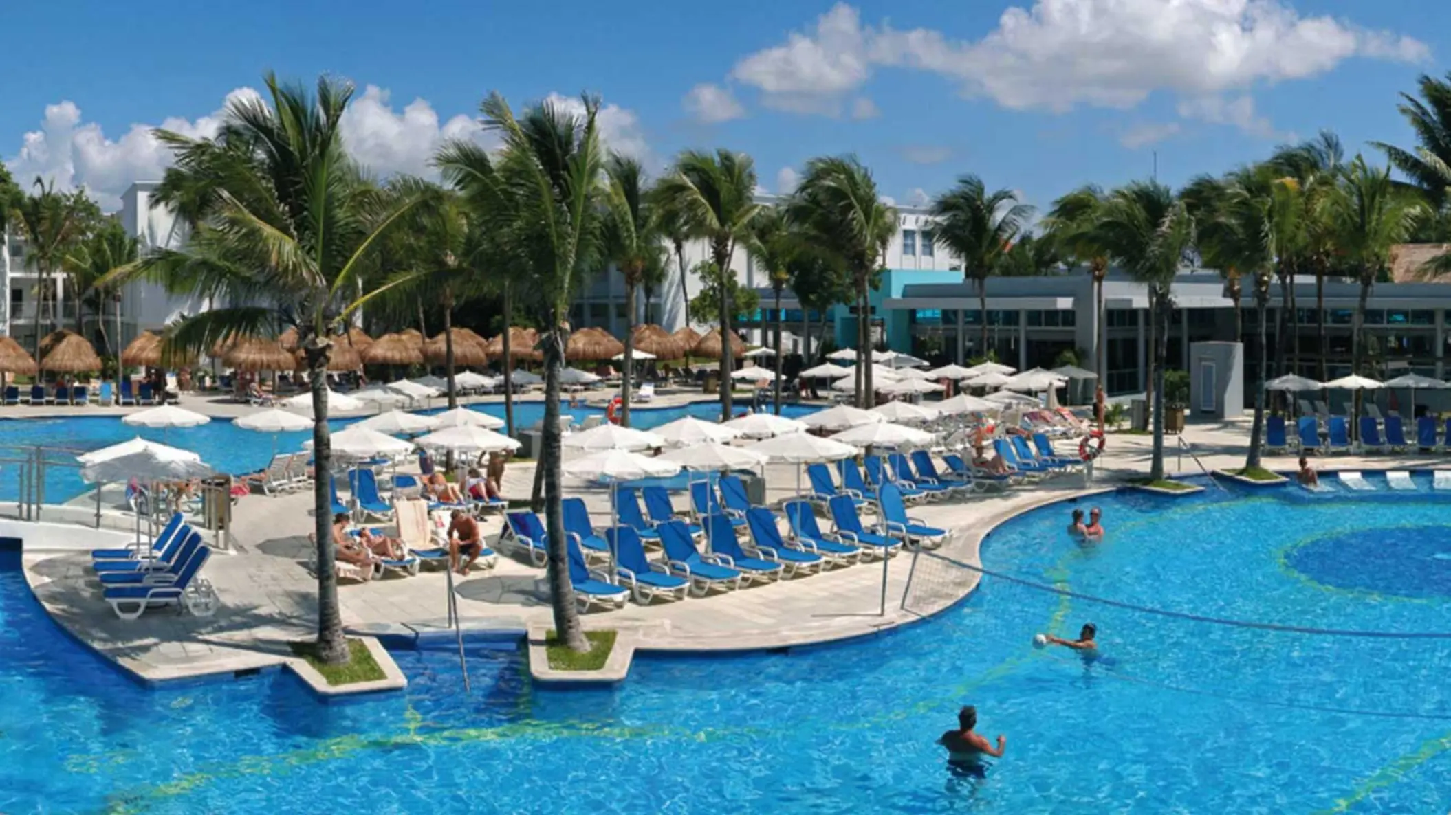 An expansive outdoor pool area at Hotel Riu Yucatan, featuring multiple interconnected pools, numerous lounge chairs, and palm trees under a bright sky.