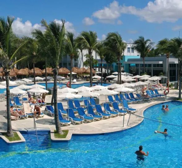 An expansive outdoor pool area at Hotel Riu Yucatan, featuring multiple interconnected pools, numerous lounge chairs, and palm trees under a bright sky.