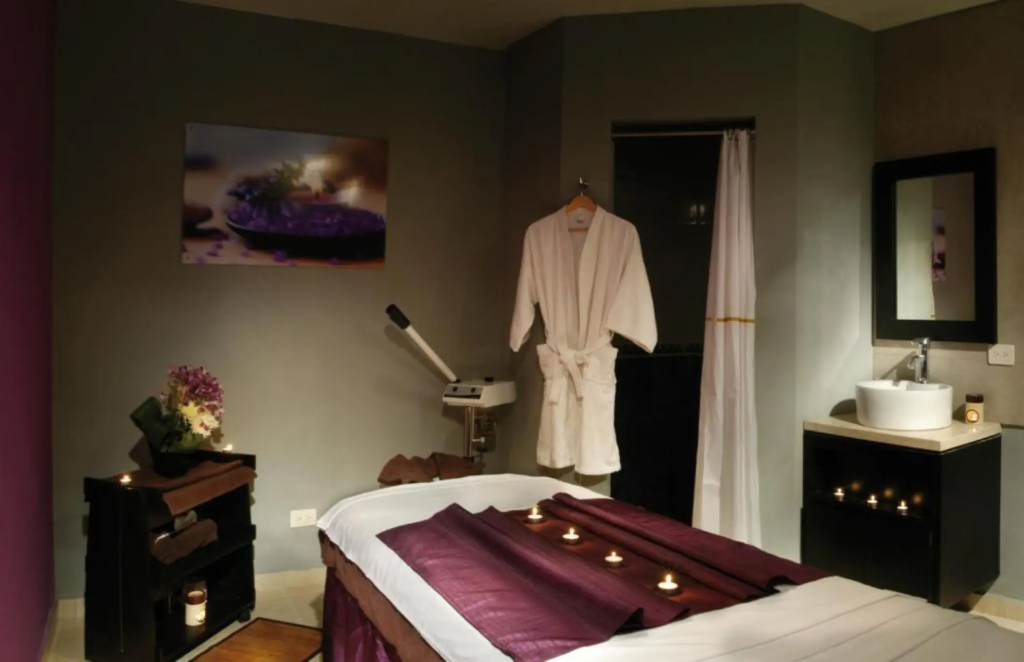 serene spa treatment room with a massage bed, a neatly folded white robe, a sink vanity, and candles for ambiance.
