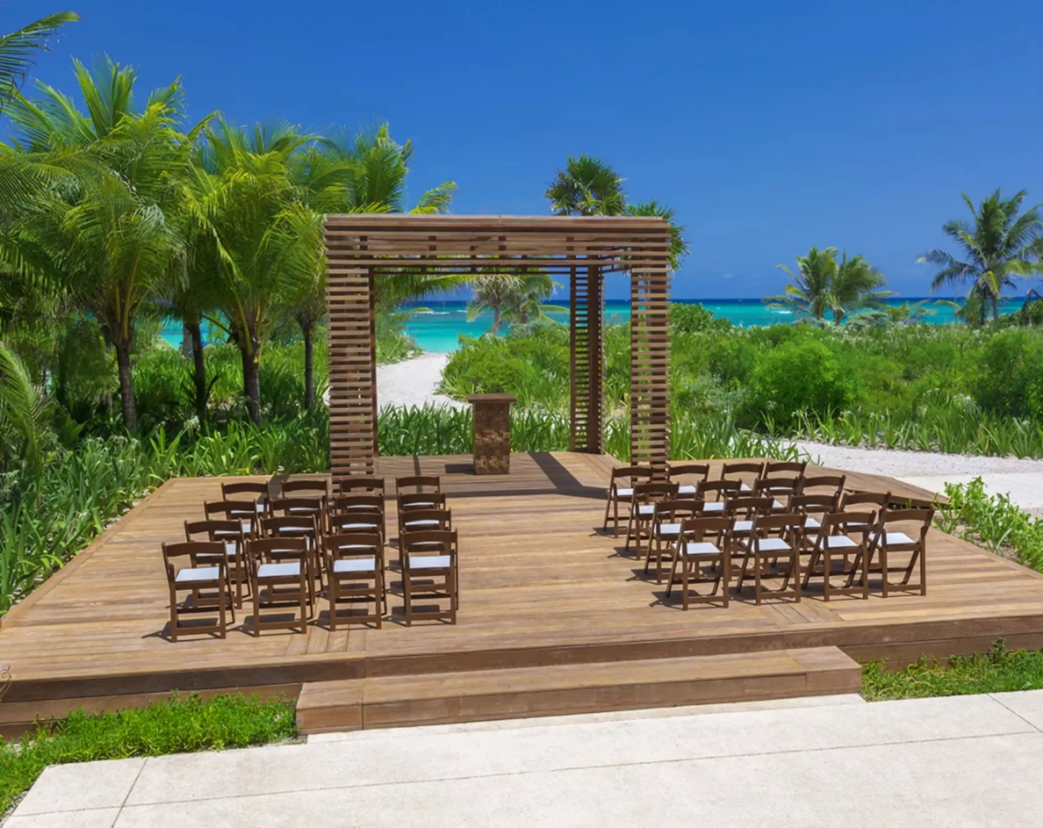Garden gazebo wedding venue with wooden platform and dark chairs, surrounded by lush palms and a view of the ocean.