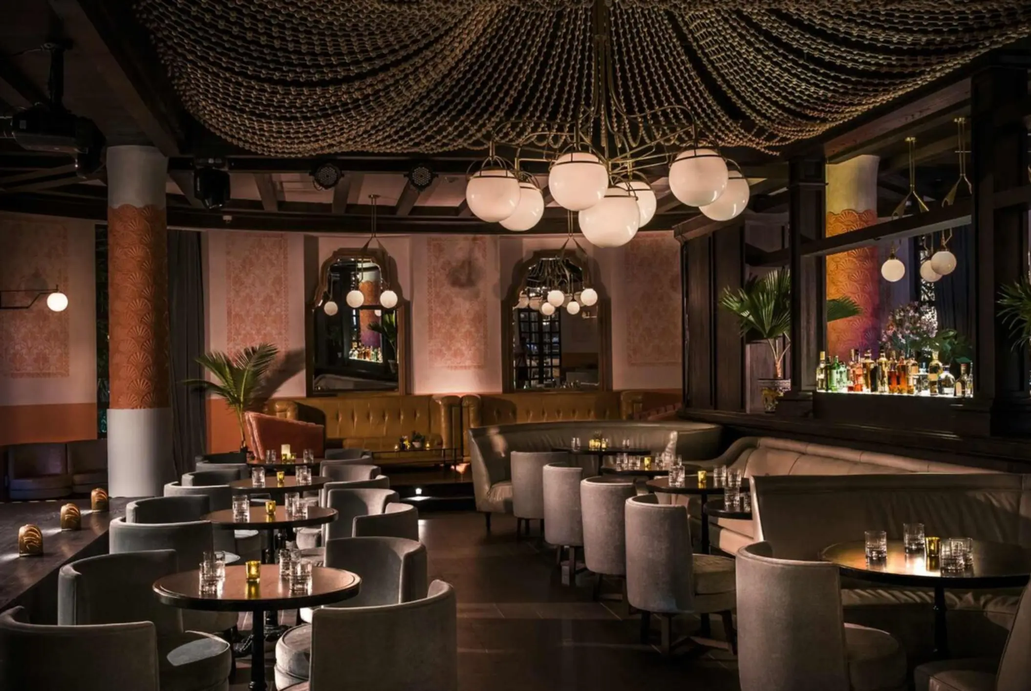 Upscale cocktail bar with plush seating, dramatic beaded ceiling fixture, and softly lit bottles behind the bar.