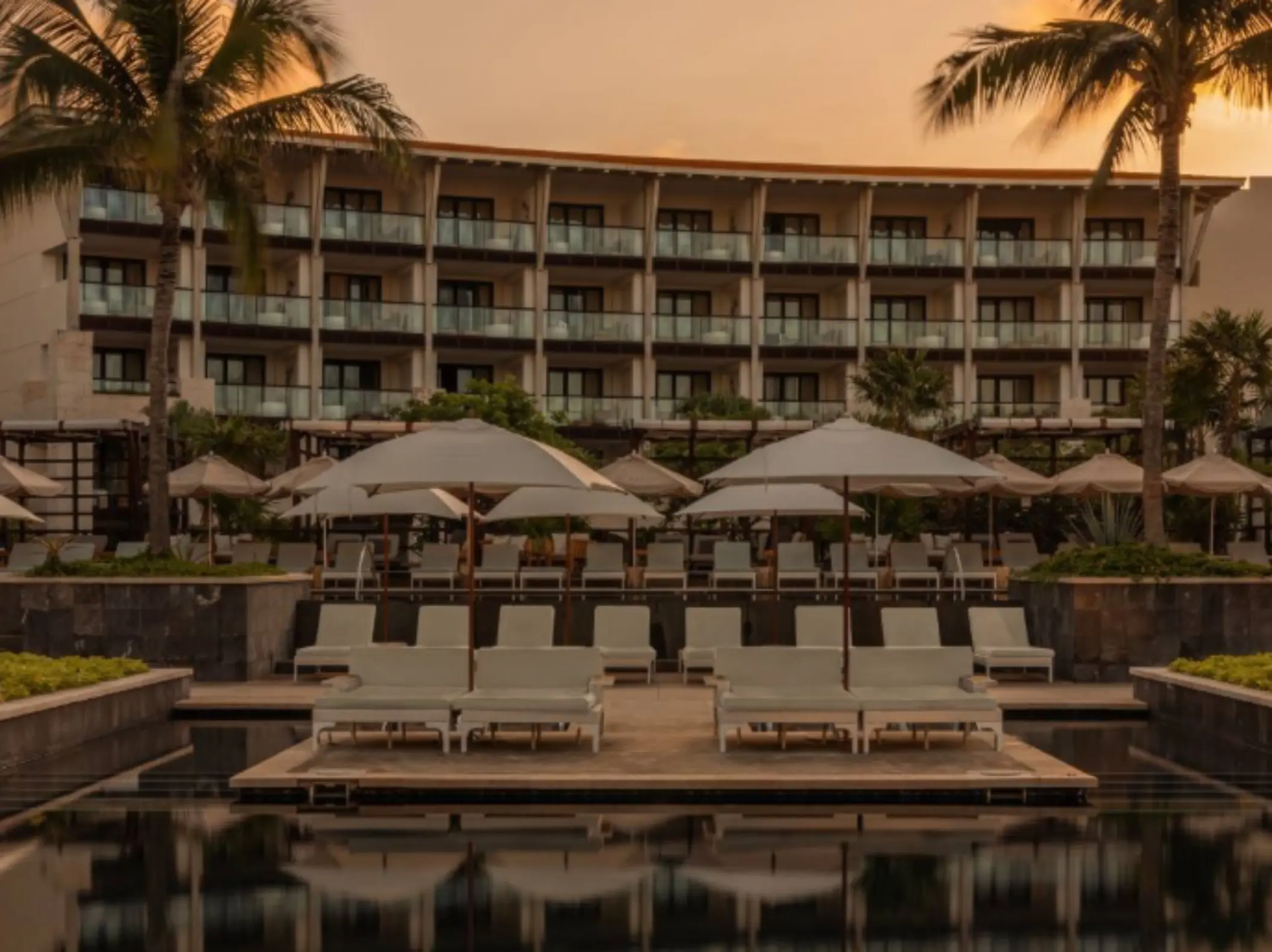 Resort pool area at sunset with overwater loungers, tall palm trees, and guest suites visible in the background.