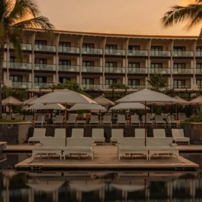 Resort pool area at sunset with overwater loungers, tall palm trees, and guest suites visible in the background.