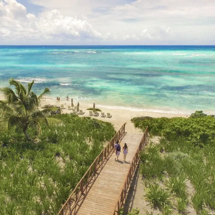 Wooden walkway through tropical greenery leading to a white sand beach with turquoise ocean views.