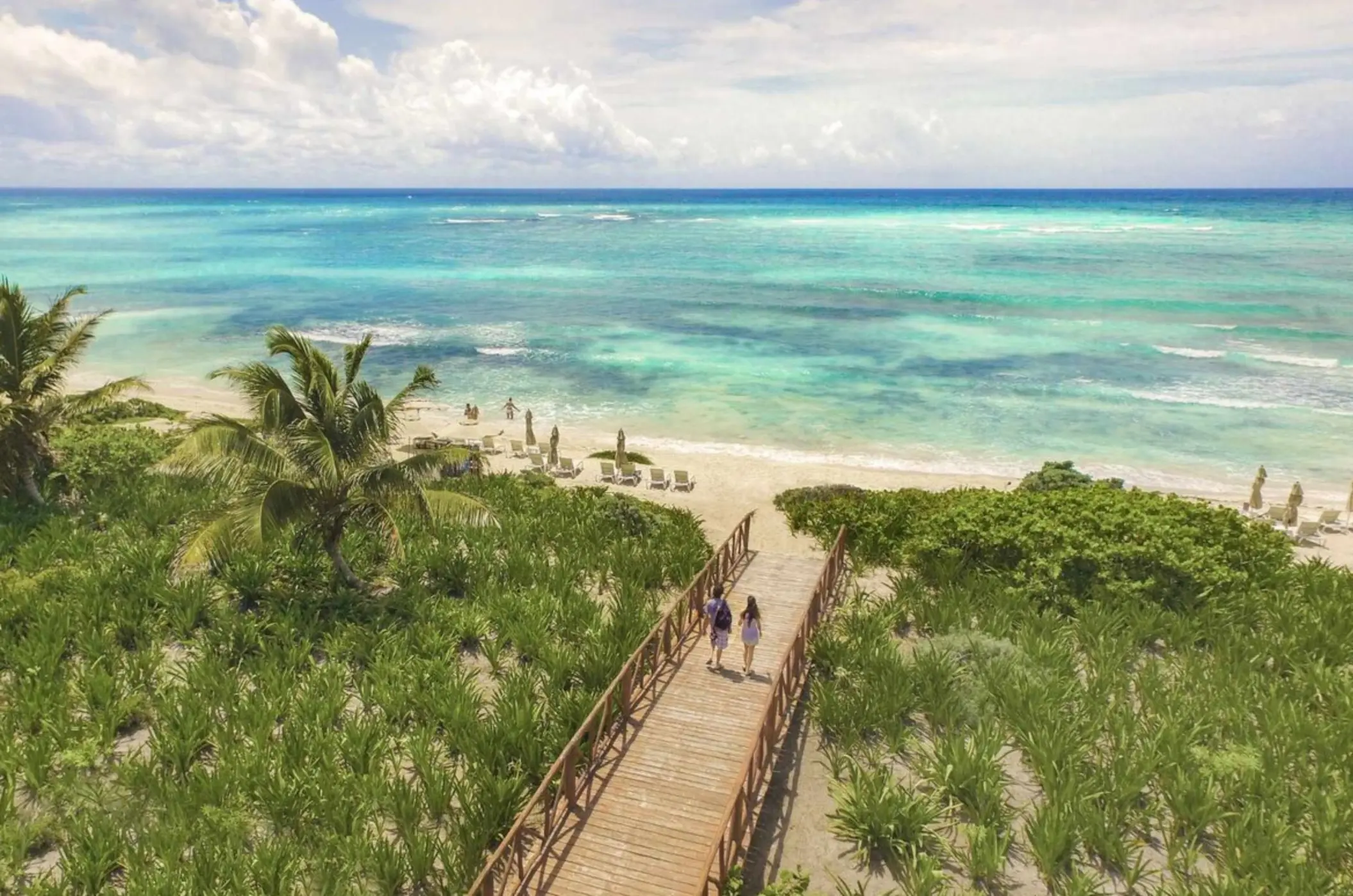 Wooden walkway through tropical greenery leading to a white sand beach with turquoise ocean views.