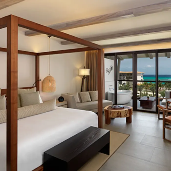Bright suite with four-poster bed, indoor seating, and ocean-view balcony with standalone soaking tub.