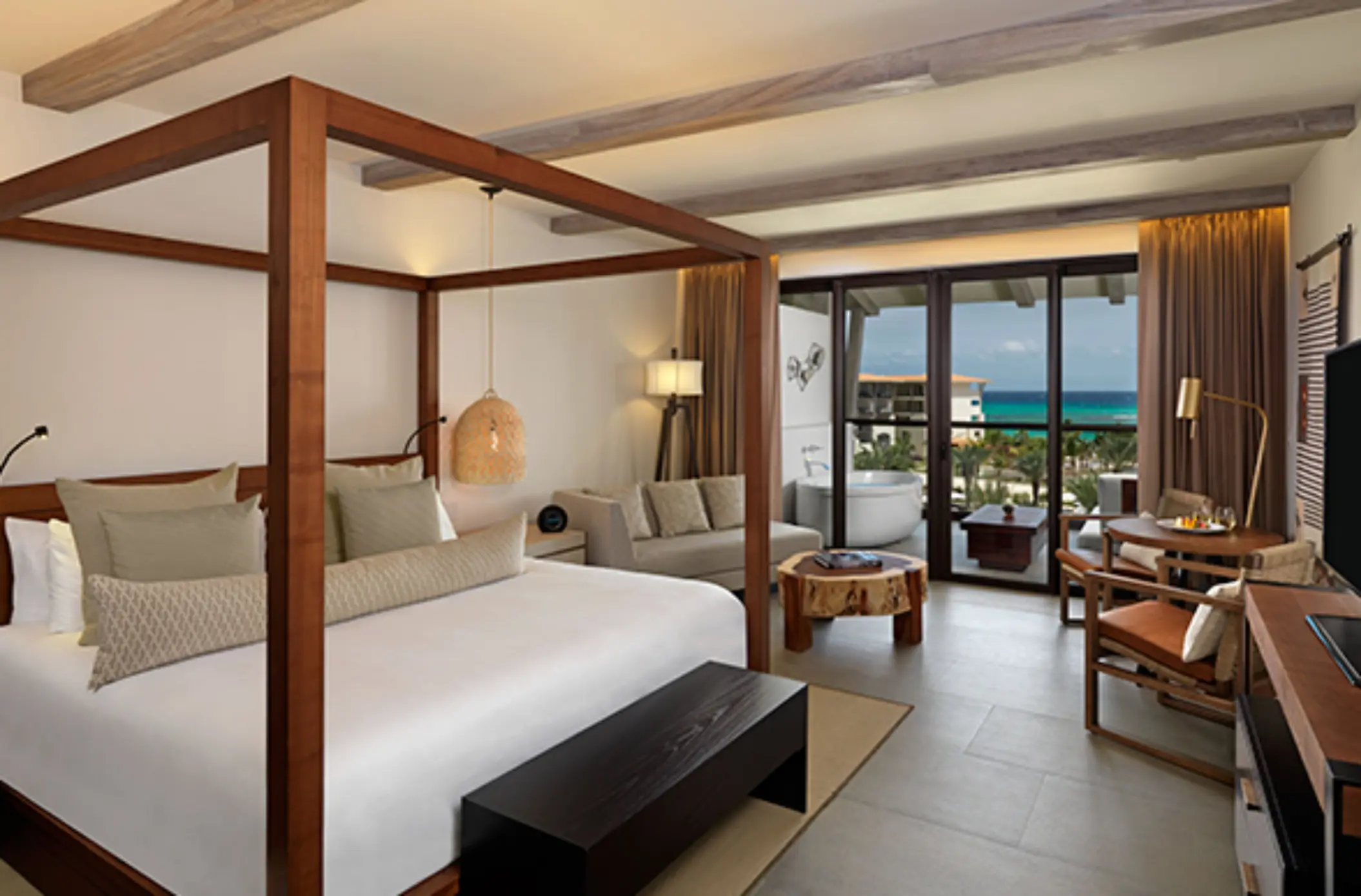 Bright suite with four-poster bed, indoor seating, and ocean-view balcony with standalone soaking tub.