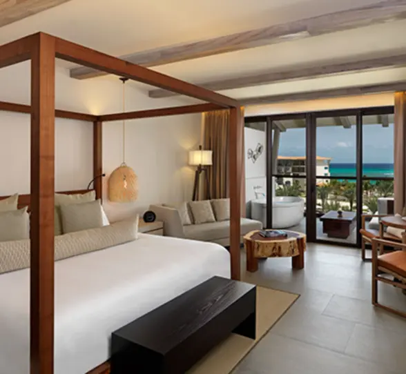 Bright suite with four-poster bed, indoor seating, and ocean-view balcony with standalone soaking tub.