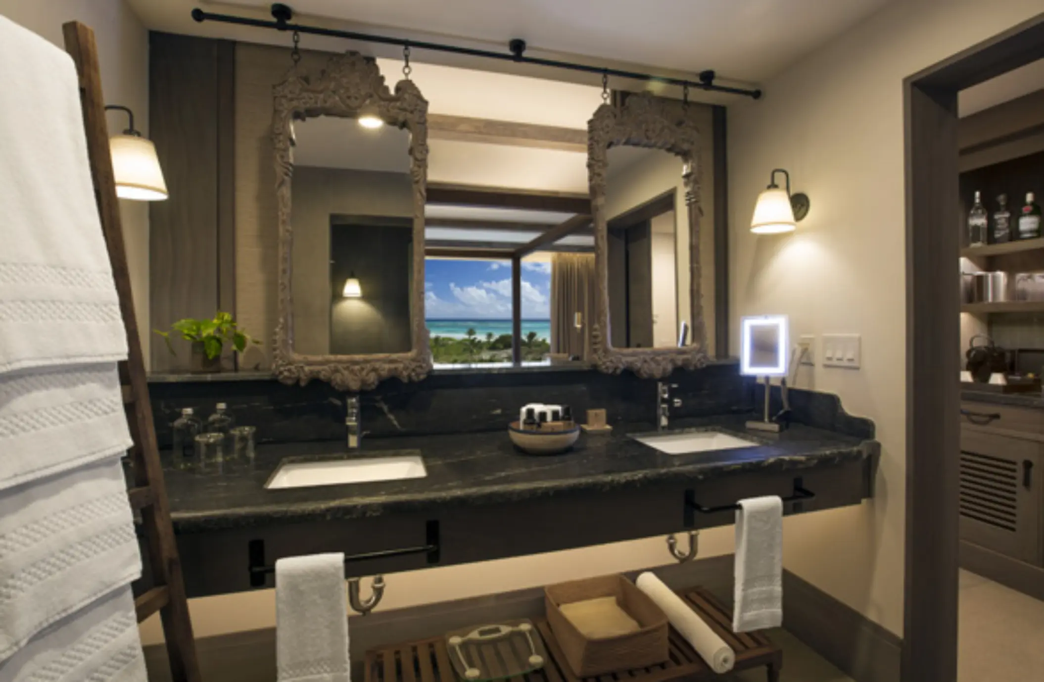 Elegant bathroom with dual sinks, framed mirrors, and window view overlooking ocean and palm trees.