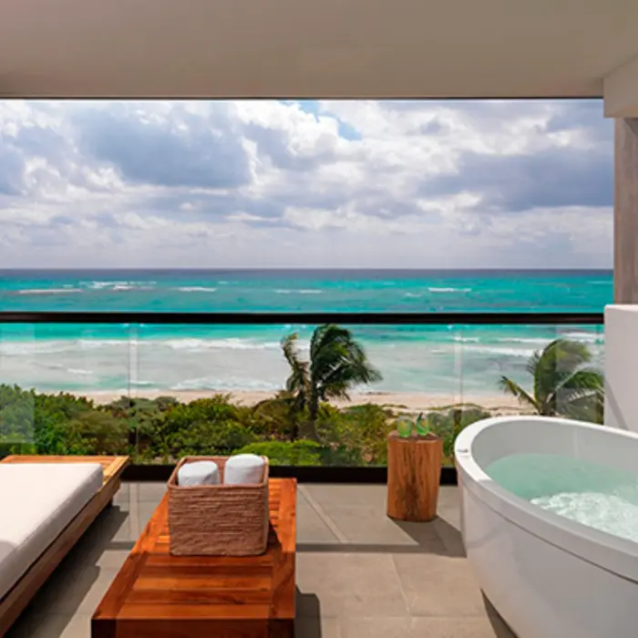 Ocean-view balcony with soaking tub, lounge bench, and wooden table overlooking turquoise waves and palm trees.