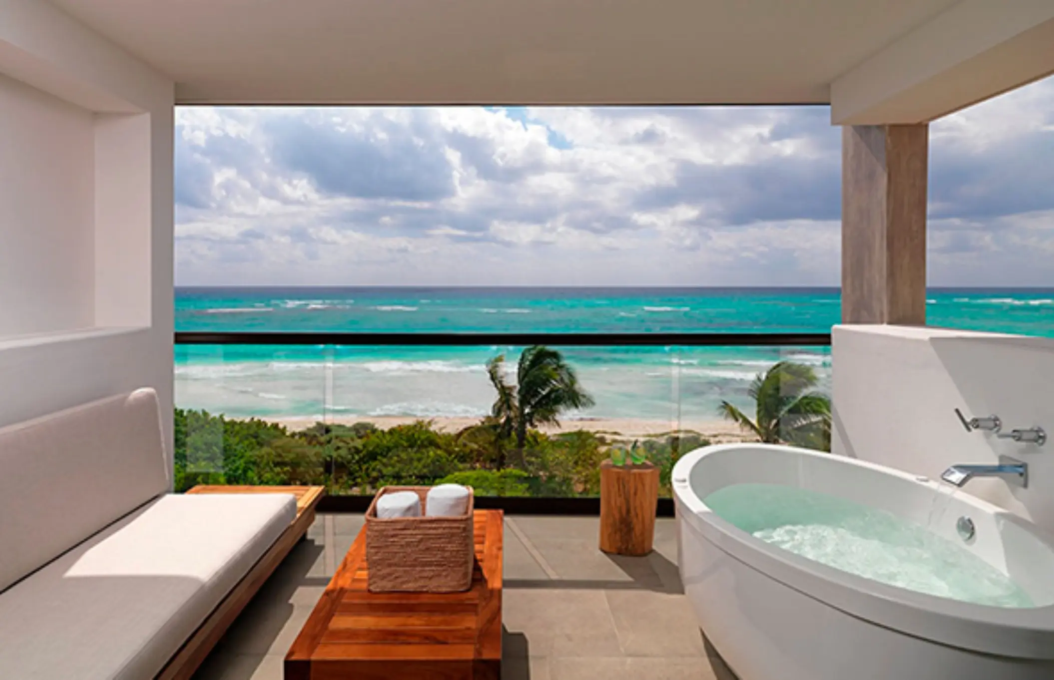 Ocean-view balcony with soaking tub, lounge bench, and wooden table overlooking turquoise waves and palm trees.