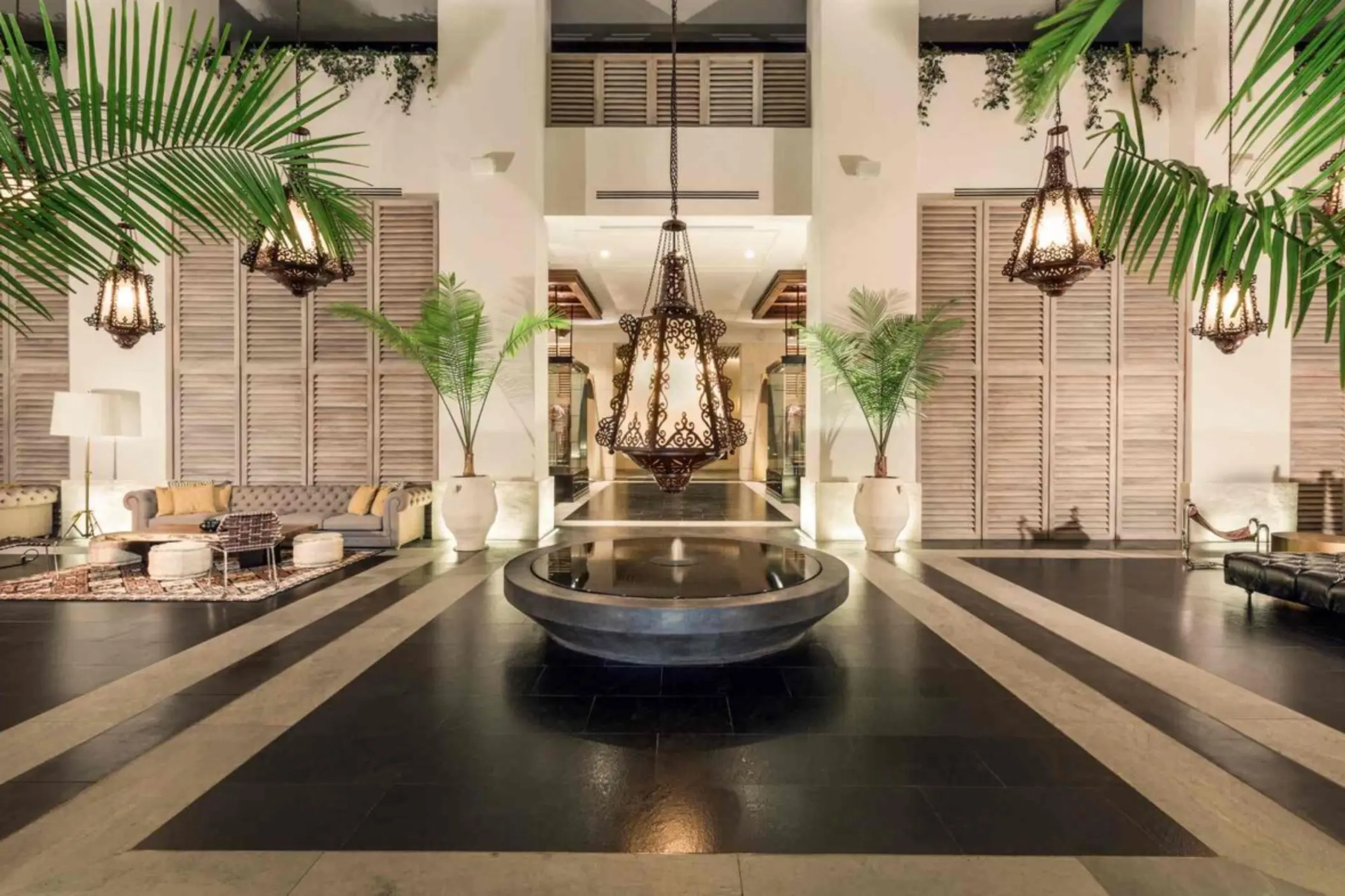 Elegant hotel lobby with Moroccan-style lanterns, modern seating, and towering potted palms flanking a fountain.