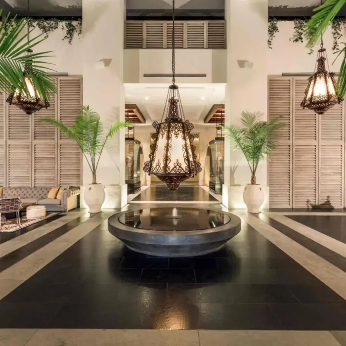 Elegant hotel lobby with Moroccan-style lanterns, modern seating, and towering potted palms flanking a fountain.