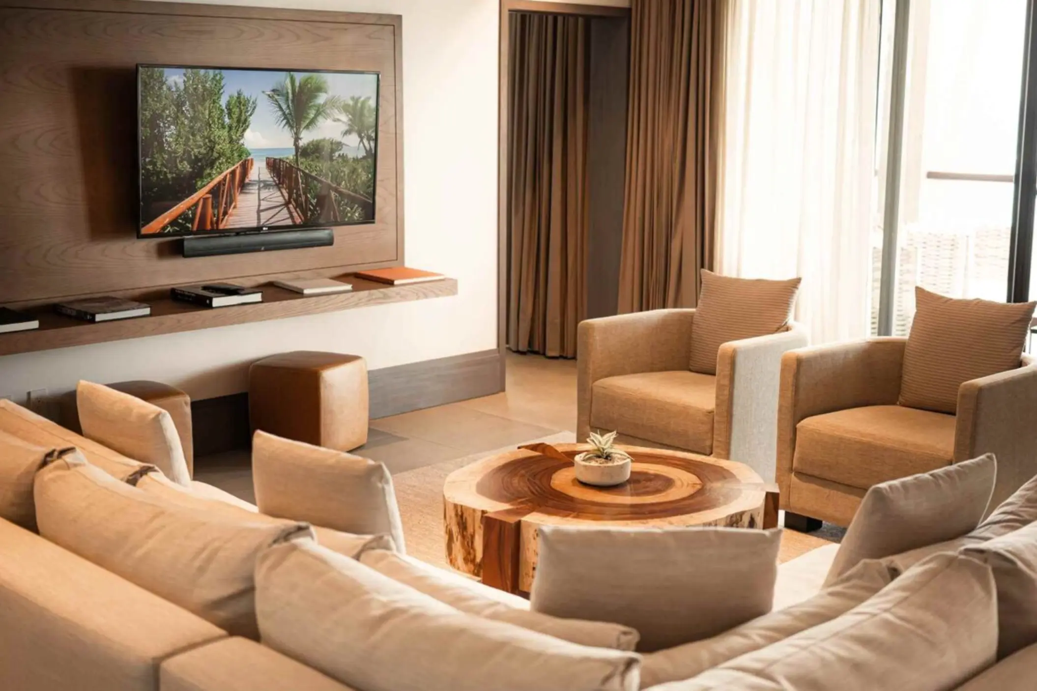 Cozy lounge area with neutral sofas, natural wood table, flat-screen TV, and sunlight pouring through the curtains.