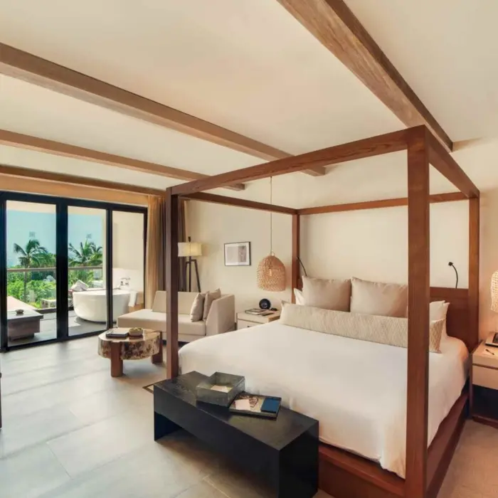 Bright and airy bedroom suite with canopy bed, seating area, and private balcony overlooking tropical greenery.