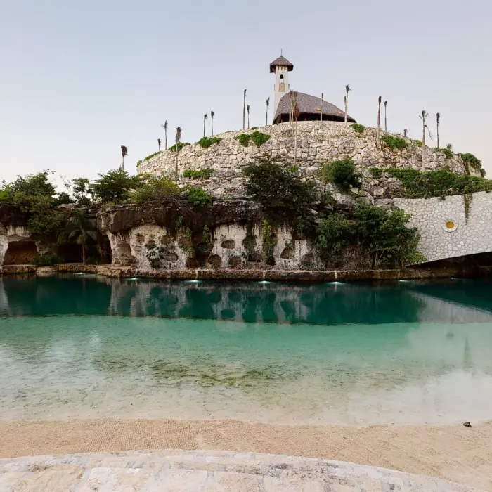 Scenic lagoon view of Hotel Xcaret Arte with a stone chapel atop a hill and lush surroundings.
