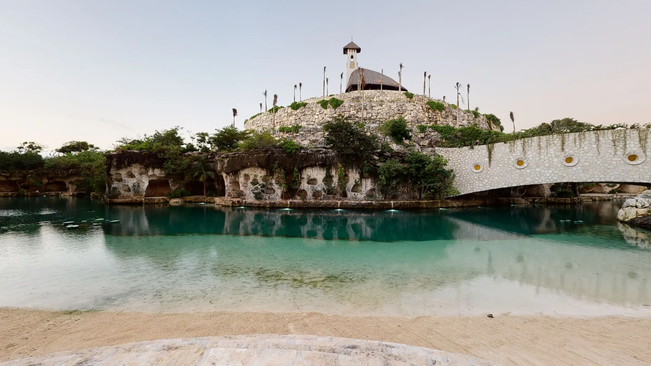 Scenic lagoon view of Hotel Xcaret Arte with a stone chapel atop a hill and lush surroundings.