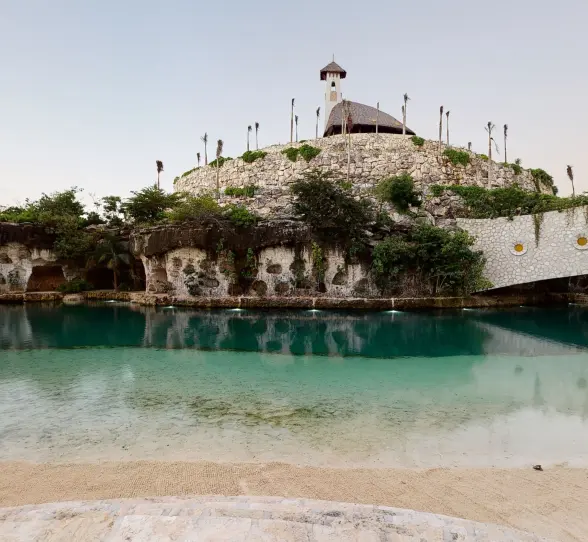 Scenic lagoon view of Hotel Xcaret Arte with a stone chapel atop a hill and lush surroundings.