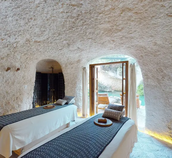 Spa cabin with massage beds inside a natural stone cave, opening to tranquil water views.