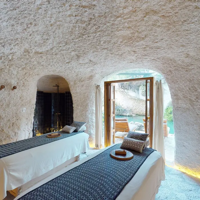 Spa cabin with massage beds inside a natural stone cave, opening to tranquil water views.