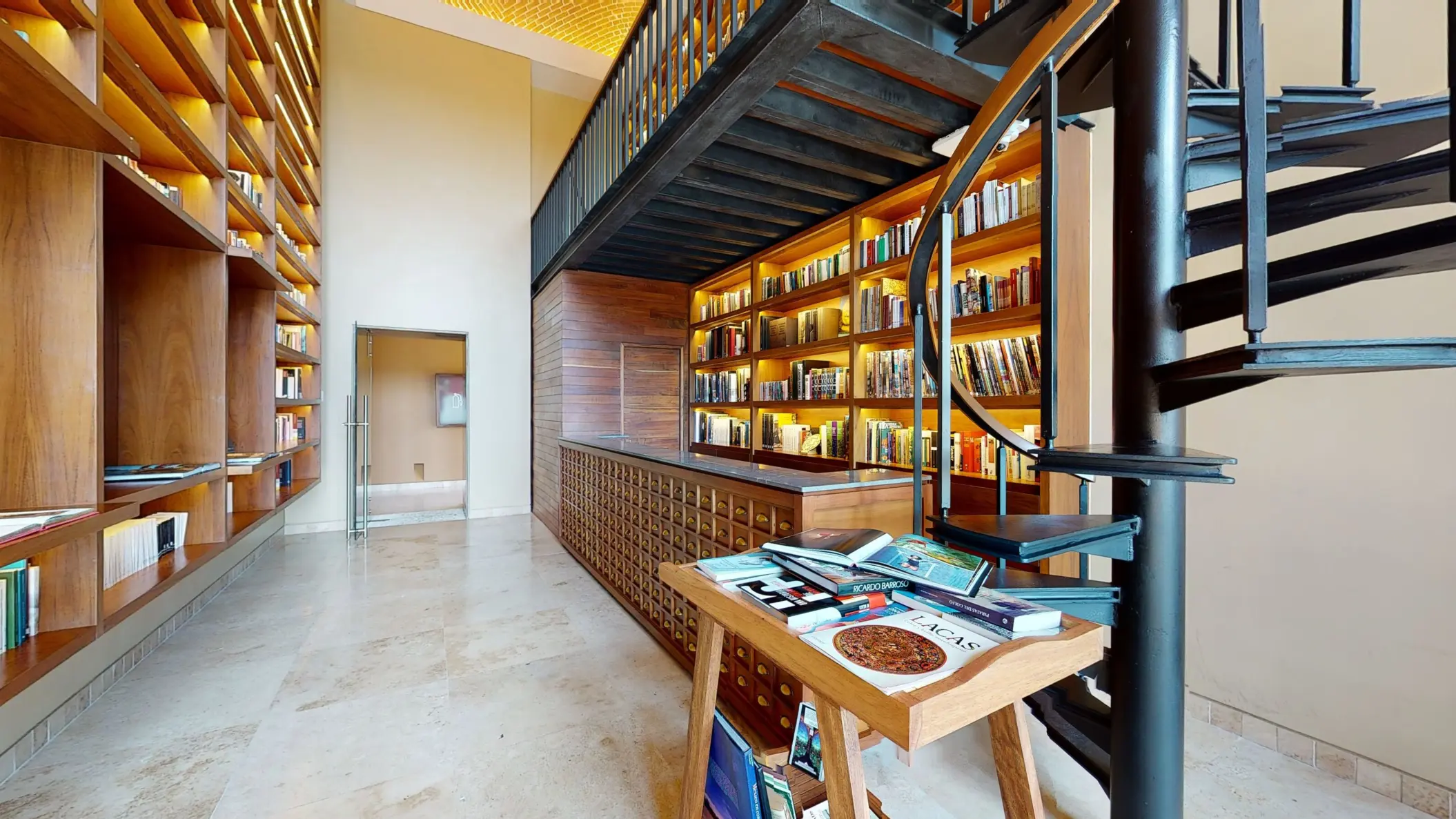 Two-level library with wood shelving, spiral staircase, and reading table filled with books and maps.