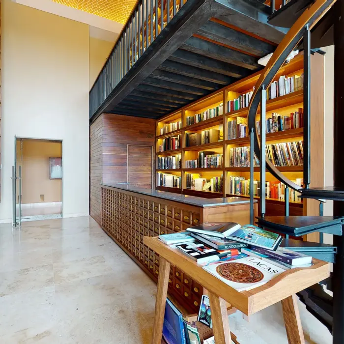 Two-level library with wood shelving, spiral staircase, and reading table filled with books and maps.