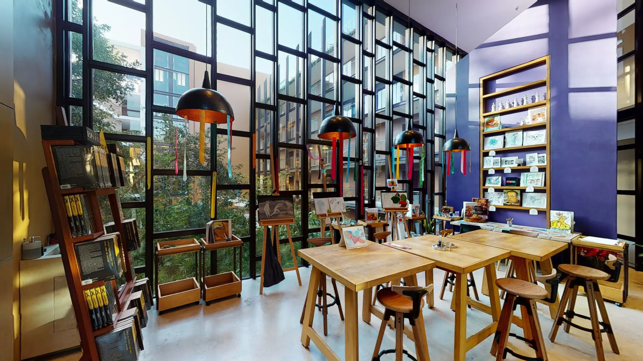Art studio with floor-to-ceiling windows, easels displaying paintings, and colorful hanging lights.