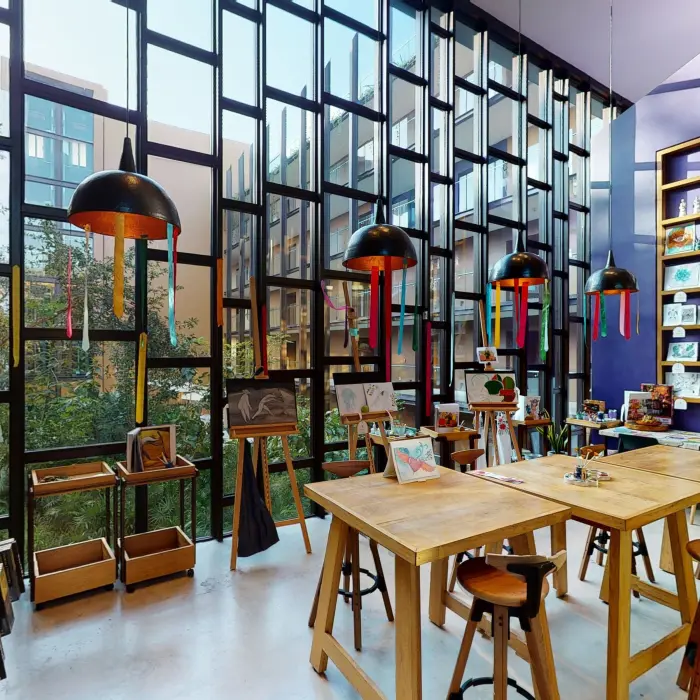 Art studio with floor-to-ceiling windows, easels displaying paintings, and colorful hanging lights.