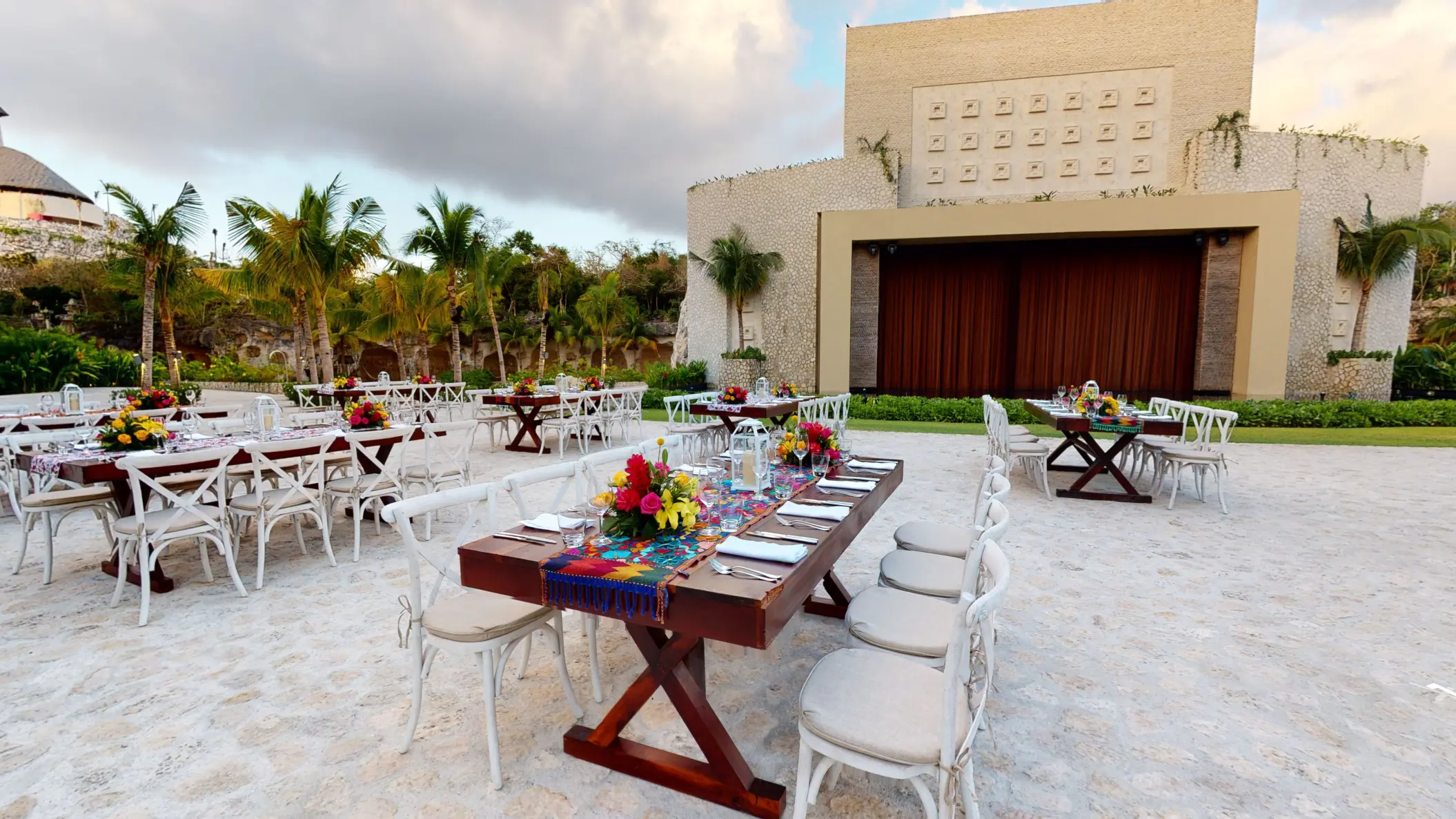 Outdoor wedding reception setup with long wooden tables, white chairs, and vibrant floral centerpieces.