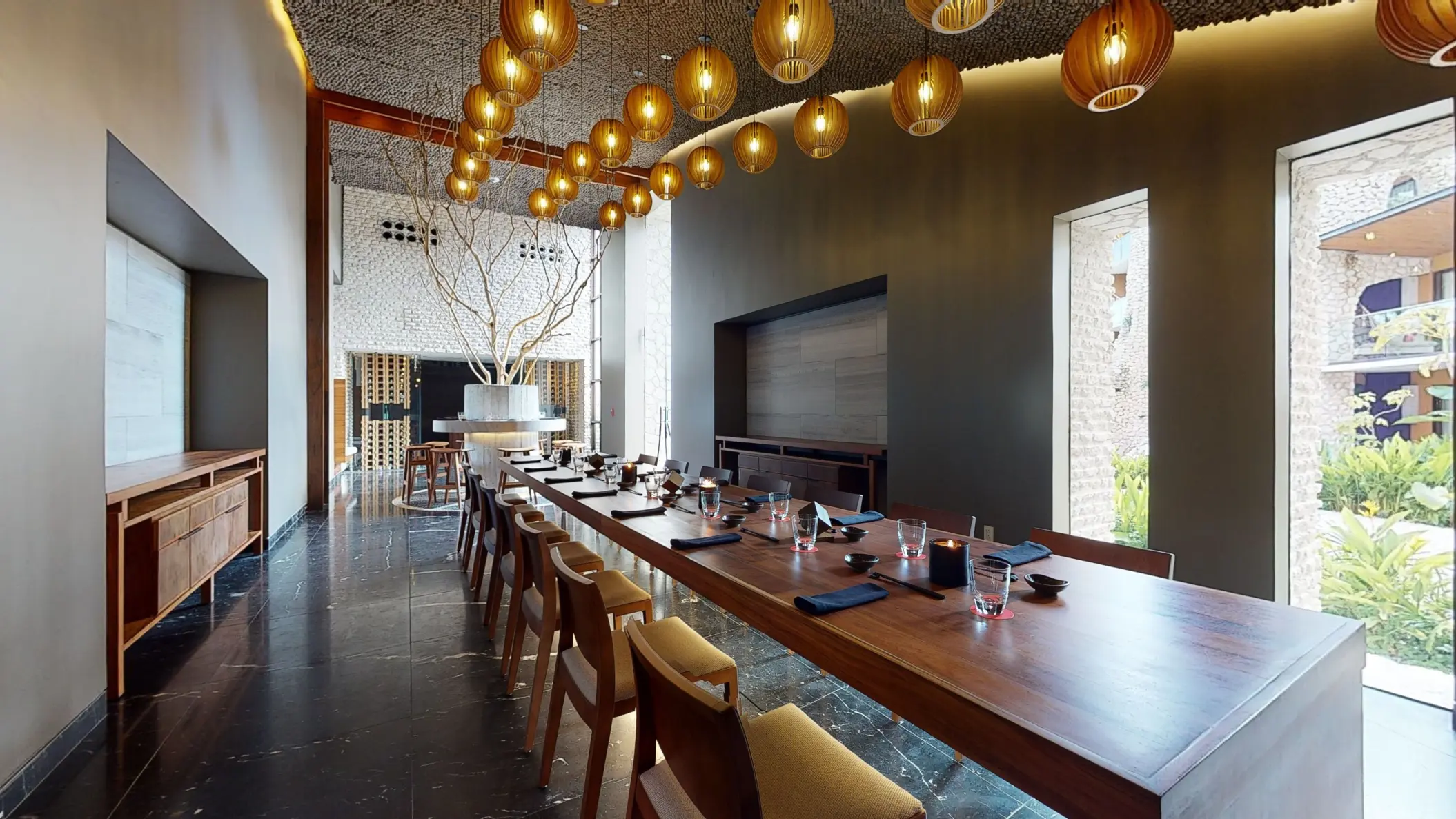 Elegant private dining room with a long wood table, pendant lights, and garden views through tall windows.