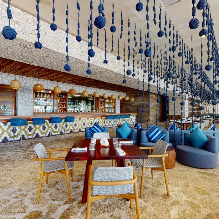 Beach-inspired bar with blue hanging ropes, cozy lounge seating, and mosaic tile bar under natural stone.