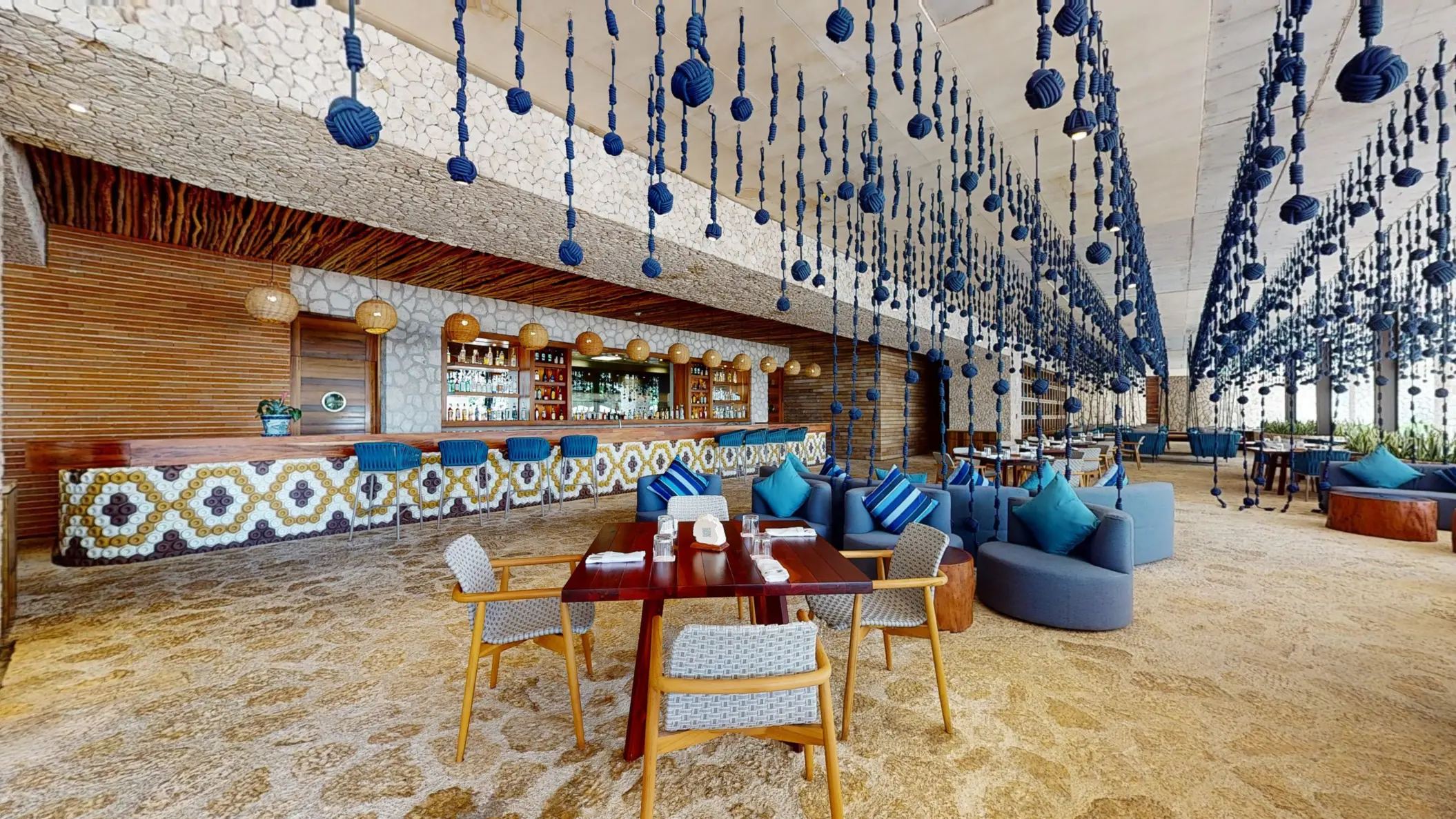 Beach-inspired bar with blue hanging ropes, cozy lounge seating, and mosaic tile bar under natural stone.