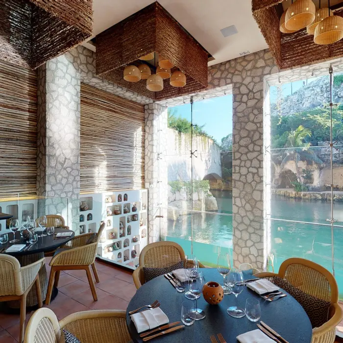 Cozy riverside restaurant with stone walls, rattan chairs, and floor-to-ceiling windows overlooking the water.