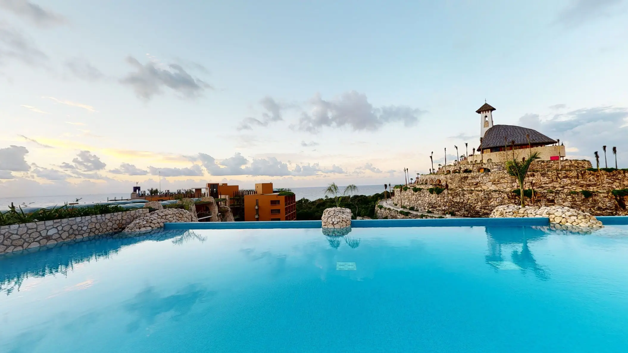 Infinity rooftop pool at sunset with panoramic ocean views and chapel perched on stone terrace.