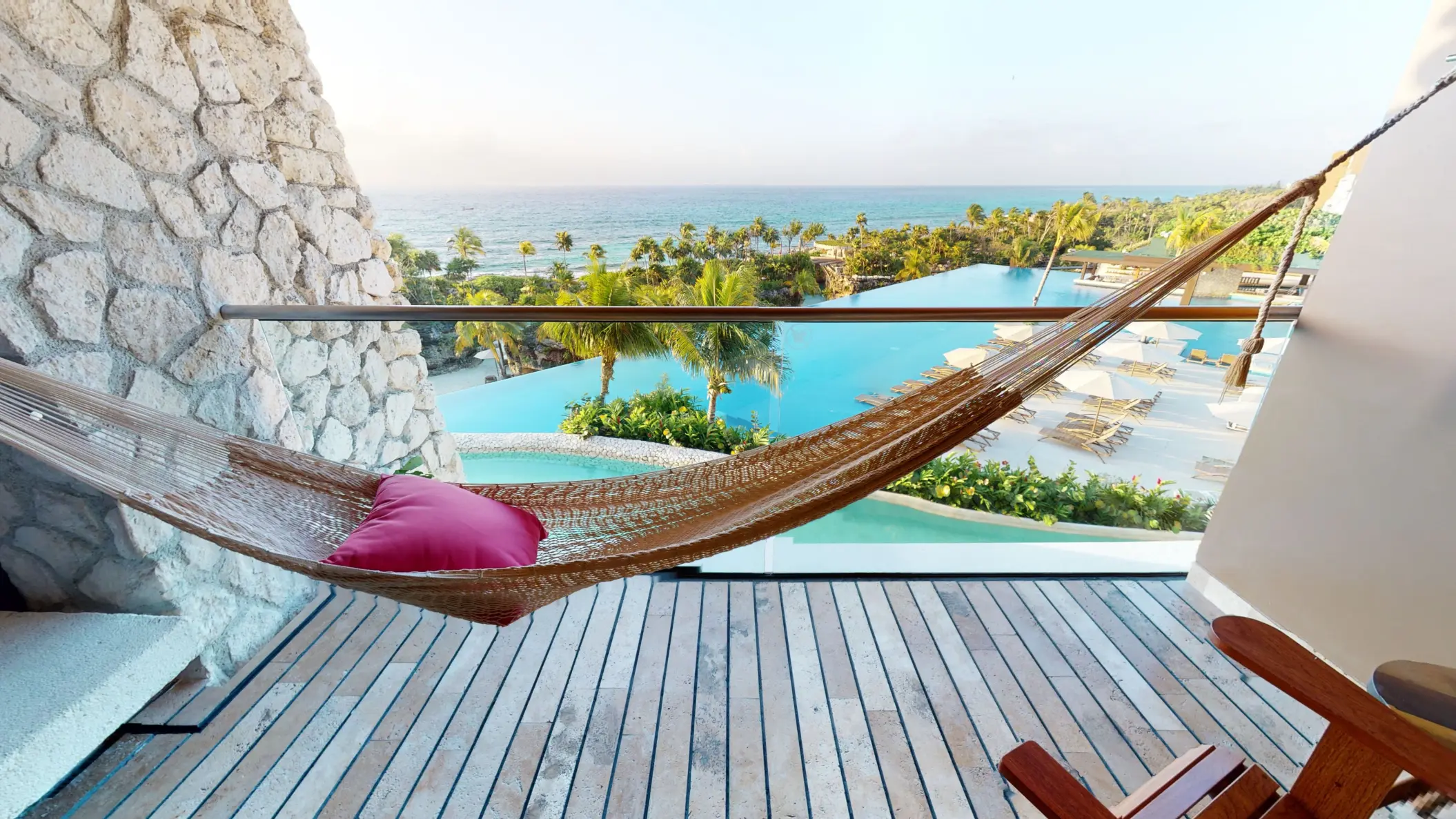 Hammock on balcony with panoramic ocean and pool views framed by lush palms and lounge chairs below.