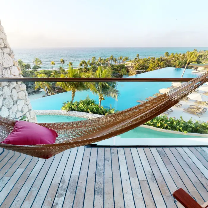 Hammock on balcony with panoramic ocean and pool views framed by lush palms and lounge chairs below.