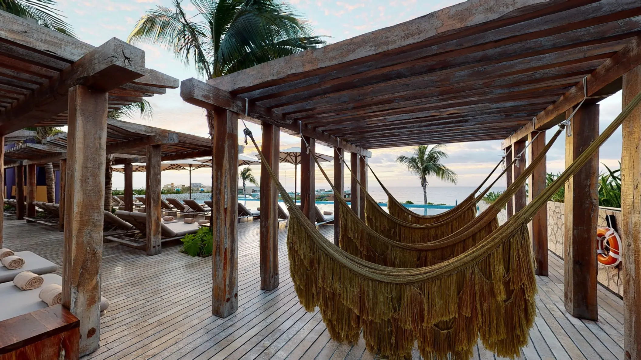 Rustic wooden pergola with hammocks and loungers facing the ocean, surrounded by palm trees.