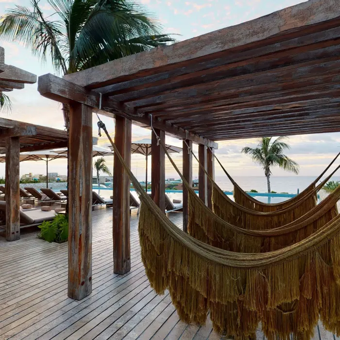 Rustic wooden pergola with hammocks and loungers facing the ocean, surrounded by palm trees.