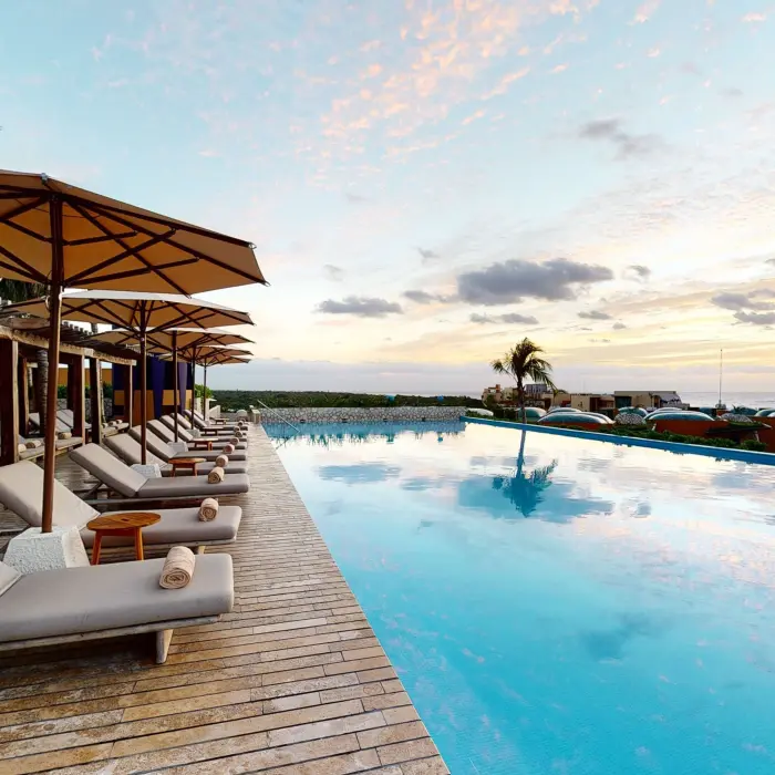 Infinity-edge pool with lounge chairs and umbrellas, overlooking the sea and tropical rooftops at sunset.